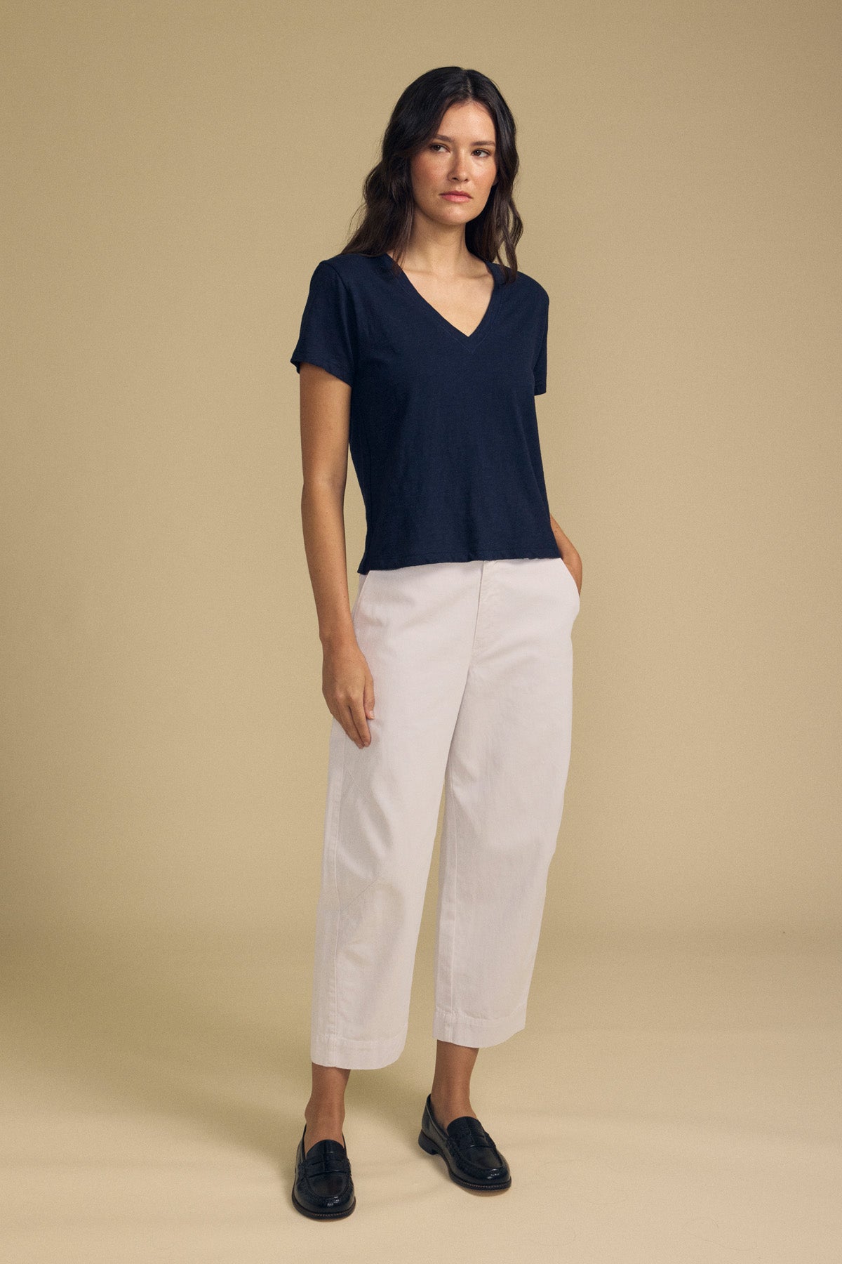 A woman stands facing forward against a plain beige background, wearing the DARIA PANT in white from Velvet by Graham & Spencer, paired with a navy V-neck T-shirt and black loafers.-41814560407745