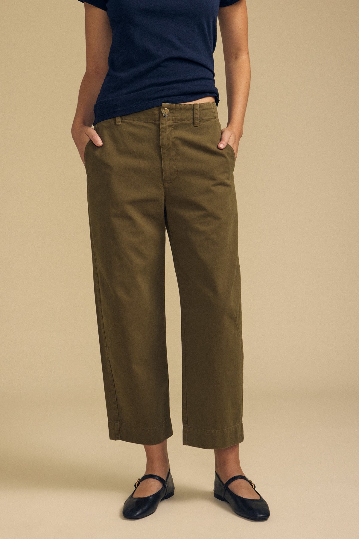 Wearing the DARIA PANT by Velvet by Graham & Spencer in olive green, paired with a navy t-shirt and black flats, this person stands with hands in pockets against a beige background.