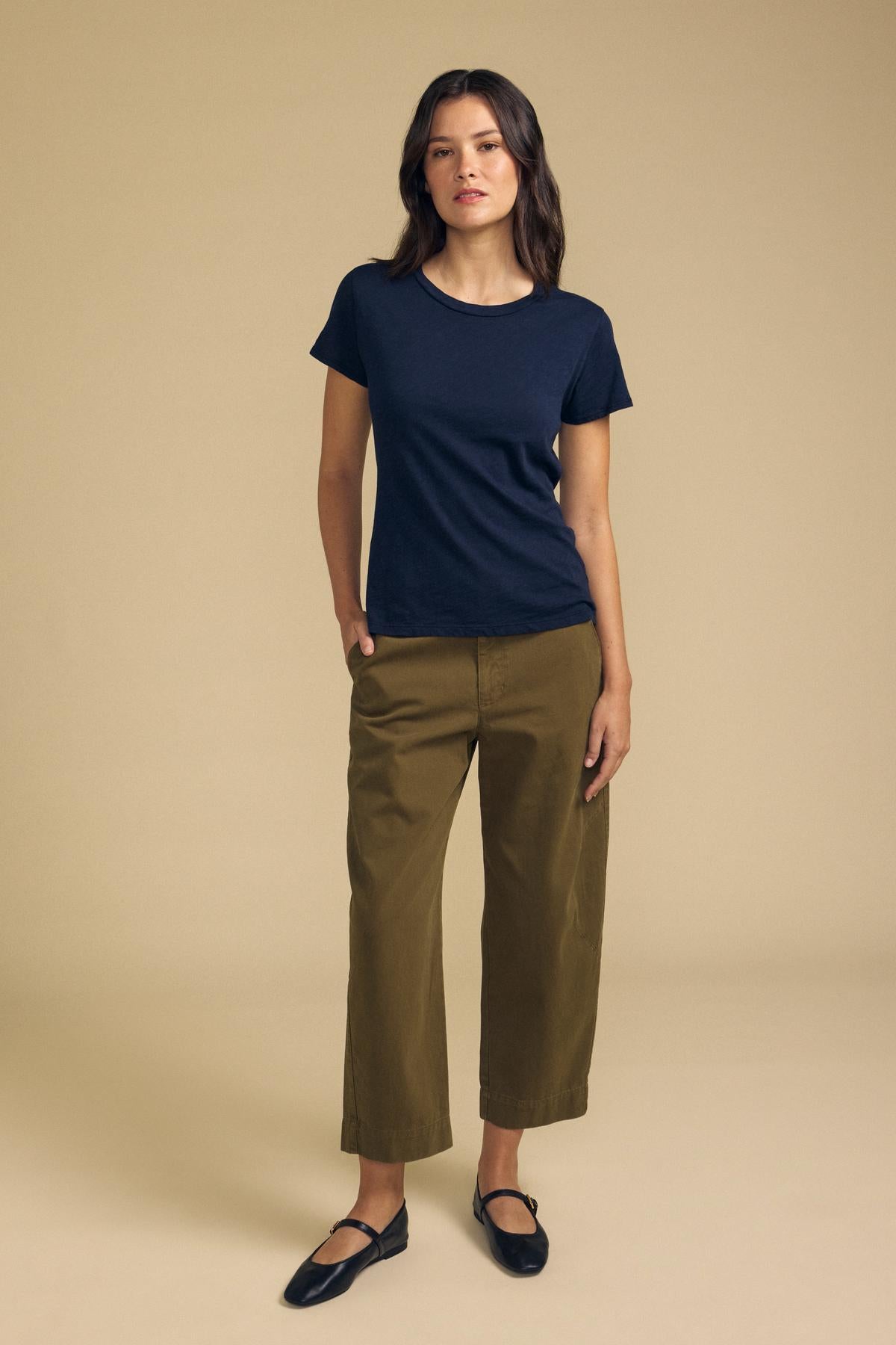 A woman stands against a beige background wearing a navy t-shirt, the DARIA PANT by Velvet by Graham & Spencer in olive green, and black flats, with one hand in her pocket.
