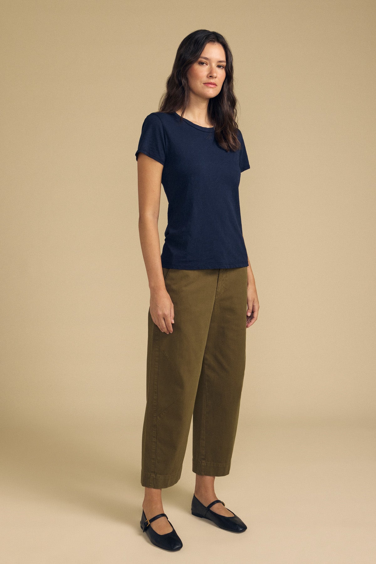 A woman stands facing forward against a plain beige background, wearing olive-green DARIA PANT by Velvet by Graham & Spencer, paired with a navy blue t-shirt and black flats.