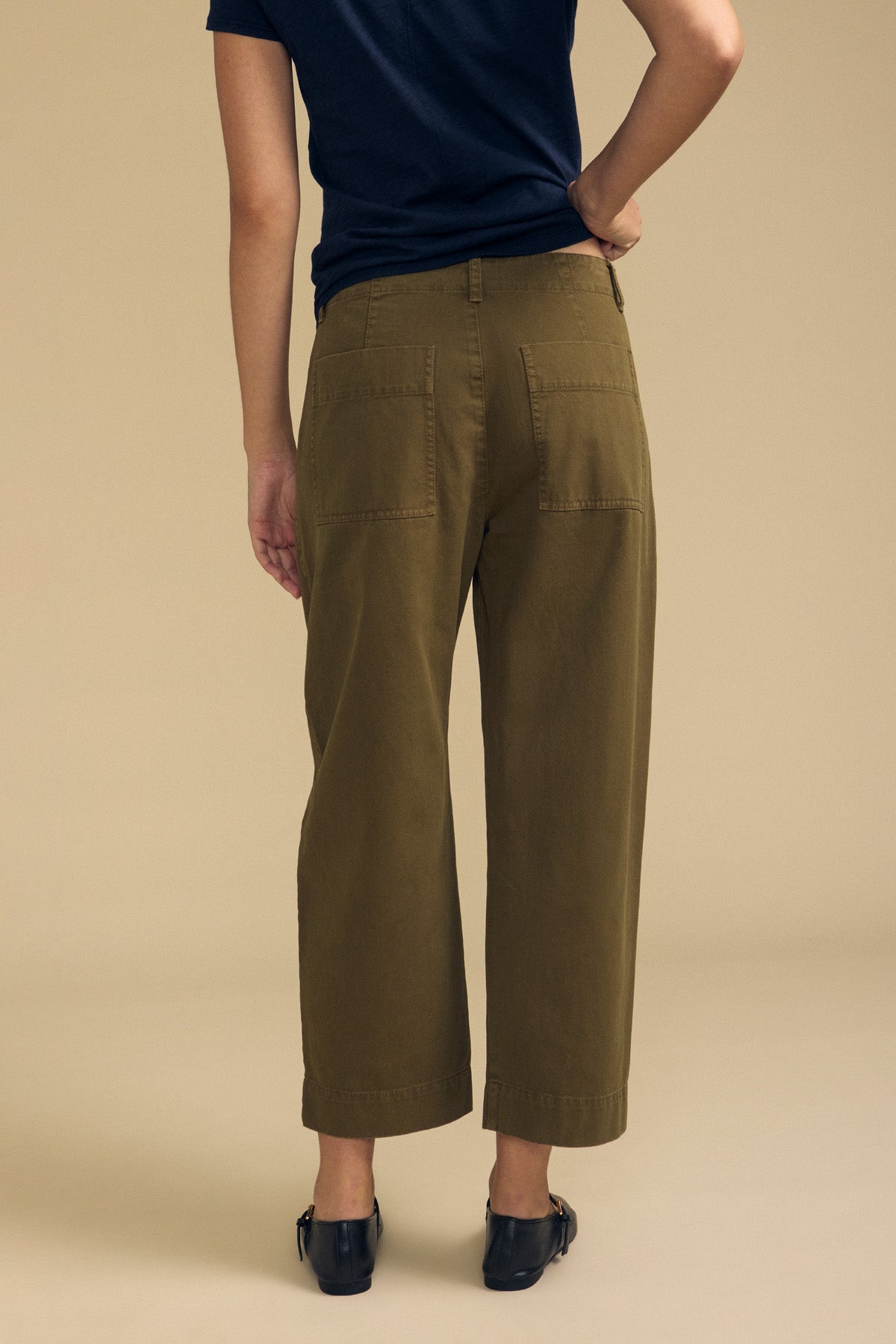 Person wearing a navy t-shirt, the DARIA PANT by Velvet by Graham & Spencer—olive green wide-leg cropped pants with back patch pockets—and black shoes, standing against a beige background.-41814560211137
