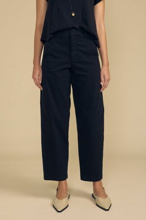 A person wearing the Velvet by Graham & Spencer TENLEY PANT—a pair of black high-waisted wide-leg pants—styled with a black top, a pendant necklace, and beige pointed-toe shoes, standing against a beige background.