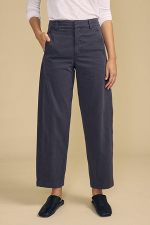A model wearing the TENLEY PANT by Velvet by Graham & Spencer—high-waisted, wide-leg, dark gray cotton twill trousers—with hands in pockets, a white long-sleeve top, and black slip-on shoes, standing against a plain beige background.
