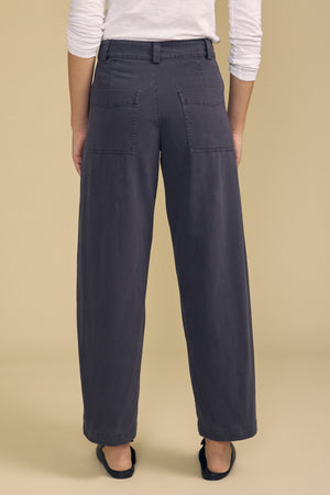 Model wearing the TENLEY PANT by Velvet by Graham & Spencer—a pair of high-waisted, wide-leg dark gray pants with back pockets—paired with black flats, photographed from the back against a beige background.