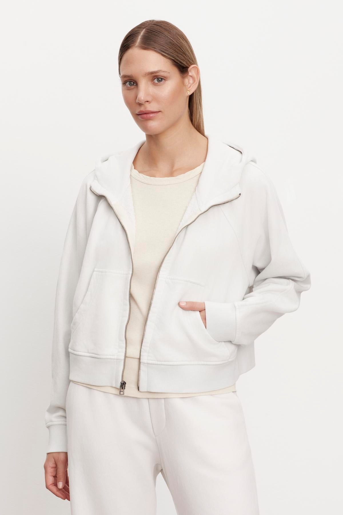 A woman stands against a plain white background wearing a Velvet by Graham & Spencer TORY SOFT FLEECE HOODIE, cream top, and white pants. She has straight brown hair and her hands in her jacket pockets.-36328606400705