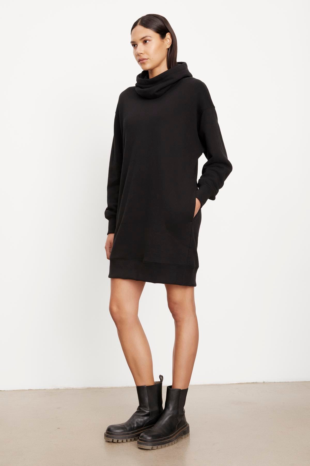   A woman in a black YARA SOFT FLEECE HOODIE DRESS by Velvet by Graham & Spencer and black boots standing against a plain background. 