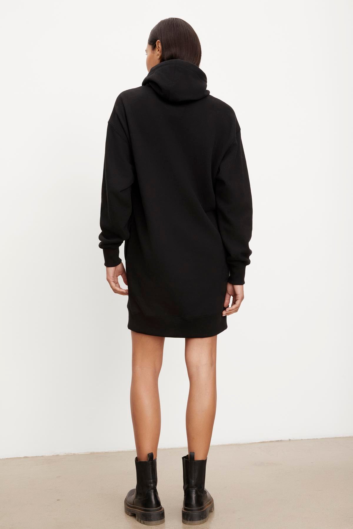 A person stands facing away from the camera, wearing a long black YARA SOFT FLEECE HOODIE DRESS by Velvet by Graham & Spencer and black boots, against a plain white background.-35696174432449