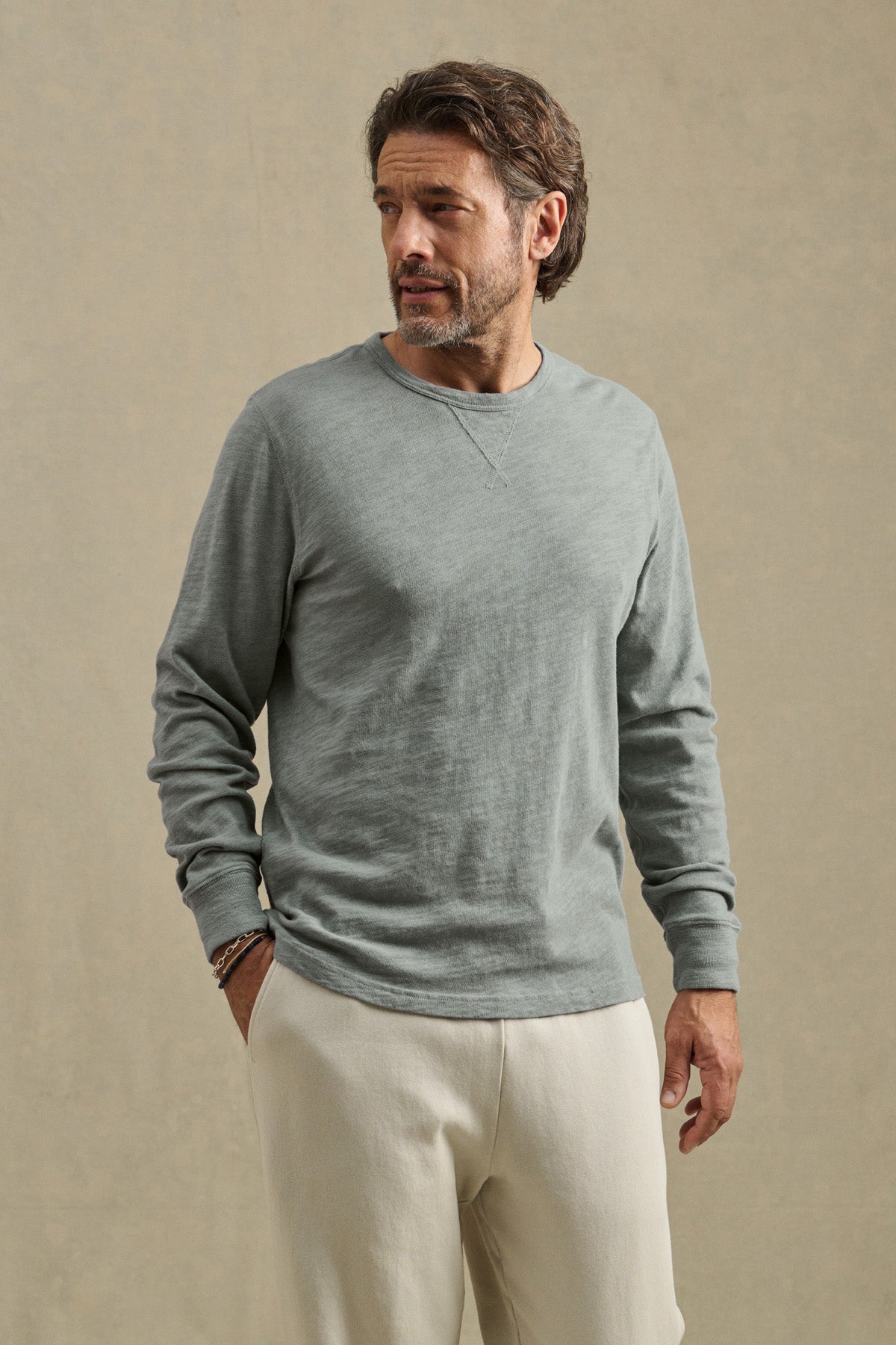 A man with a beard wears the Velvet by Graham & Spencer DUSTIN TEE, a light gray relaxed-fit long-sleeve top, paired with light pants. He stands against a neutral background, looking to his left.
