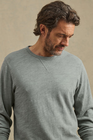 A man with brown hair and a beard wears the DUSTIN TEE by Velvet by Graham & Spencer, a relaxed fit light gray cotton slub shirt, as he looks downward against a plain beige background.