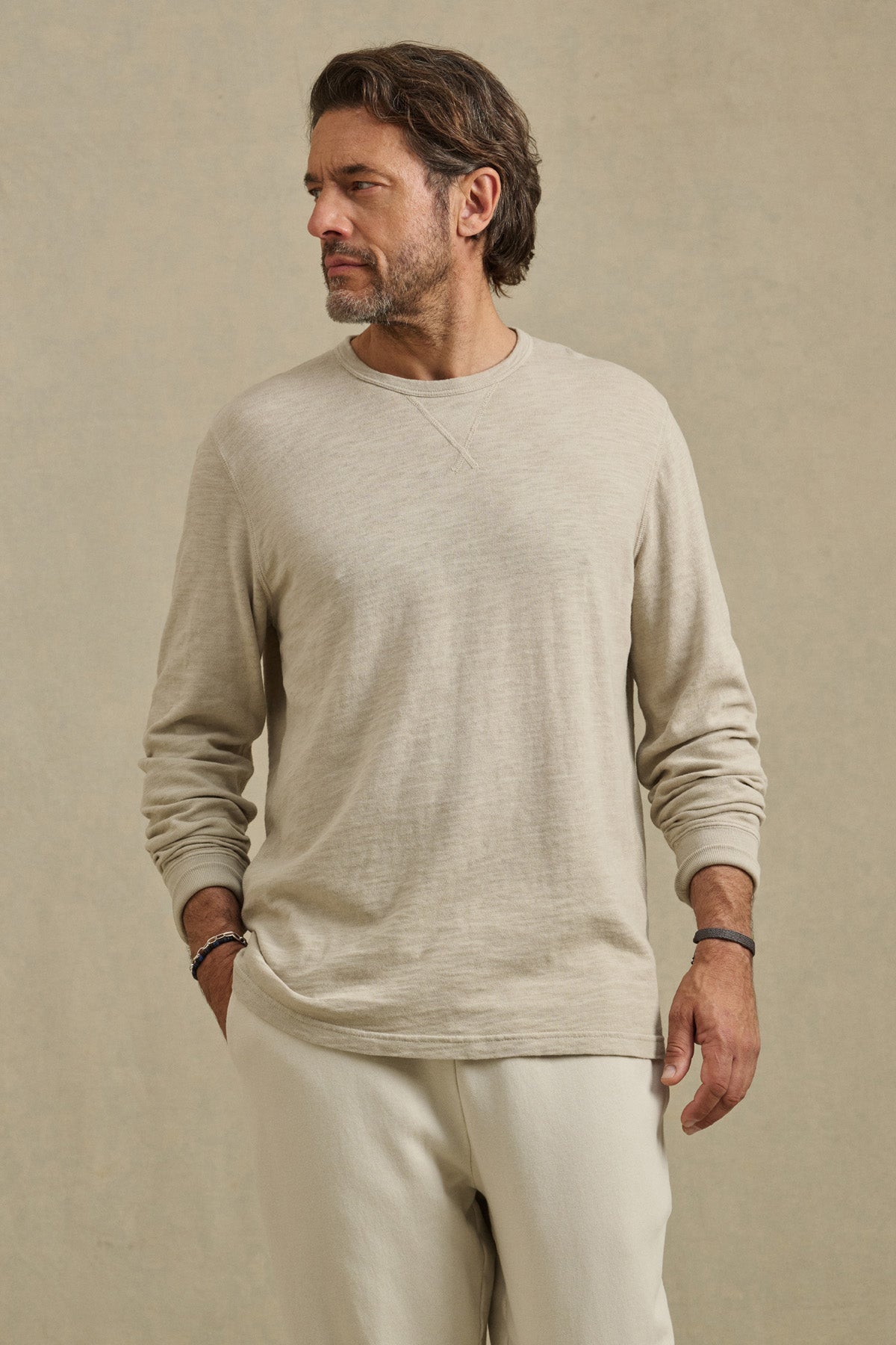 A man with short brown hair and a beard wears the Velvet by Graham & Spencer DUSTIN TEE, a relaxed fit cotton slub long-sleeve, paired with cream pants. He stands against a plain beige background, looking to his left.-41104736354497