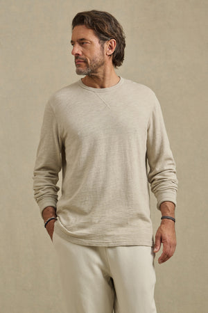 A man with short brown hair and a beard wears the Velvet by Graham & Spencer DUSTIN TEE, a relaxed fit cotton slub long-sleeve, paired with cream pants. He stands against a plain beige background, looking to his left.