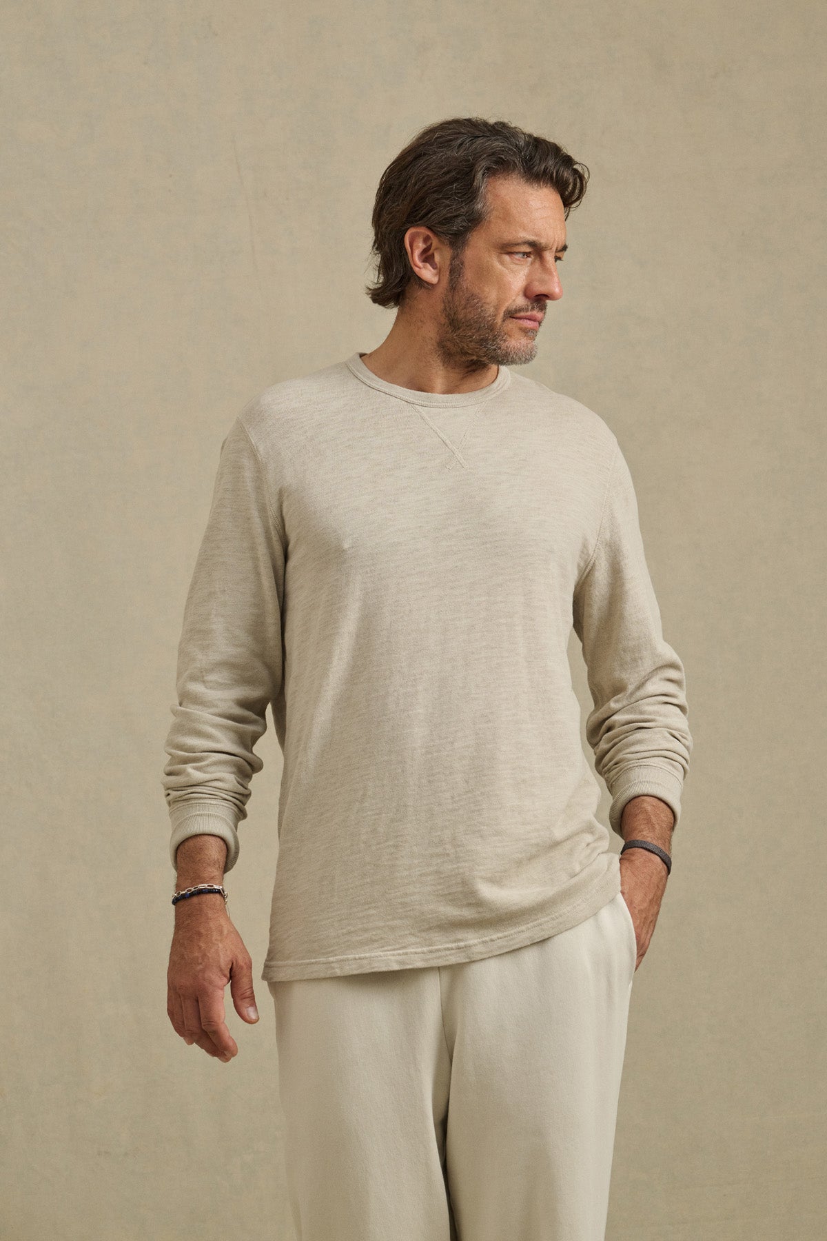 A man wearing the DUSTIN TEE by Velvet by Graham & Spencer in relaxed, light beige cotton slub stands against a plain beige background, looking to his right, with matching pants.
