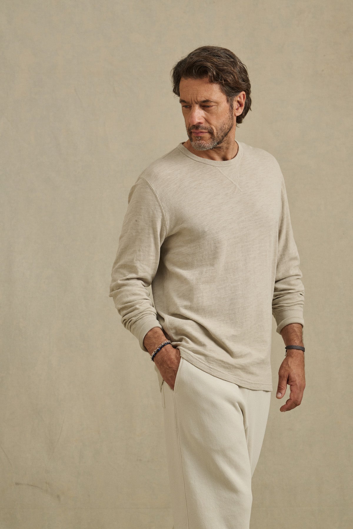 A man with a beard wears the Velvet by Graham & Spencer DUSTIN TEE, a beige cotton slub long-sleeve, and cream pants as he stands against a plain background, looking down to his left.-41104736288961