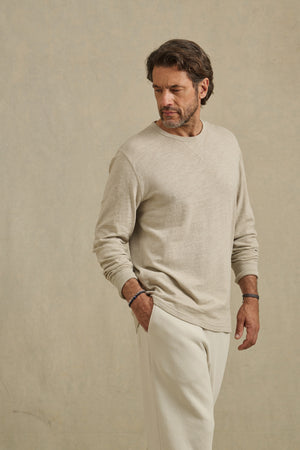 A man with a beard wears the Velvet by Graham & Spencer DUSTIN TEE, a beige cotton slub long-sleeve, and cream pants as he stands against a plain background, looking down to his left.