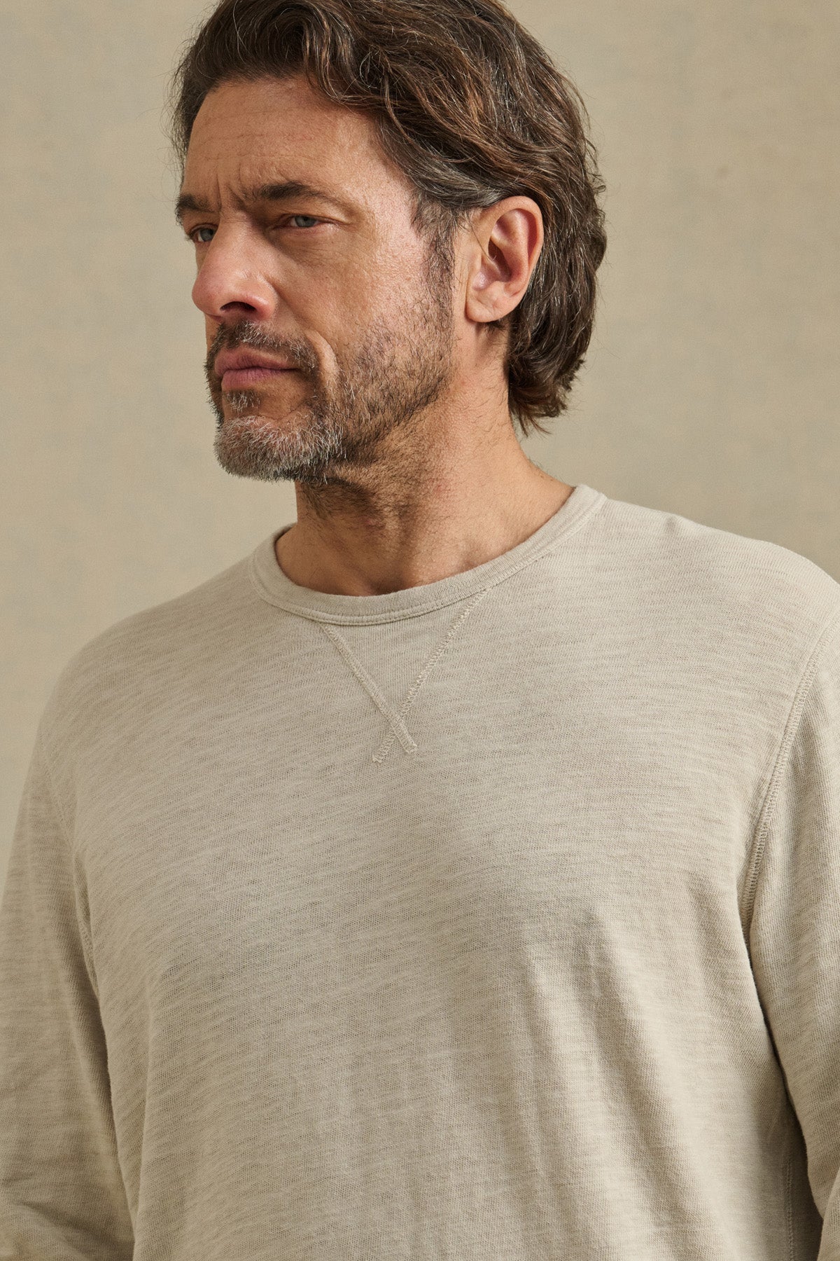 Man with medium-length brown hair and a beard wears the DUSTIN TEE by Velvet by Graham & Spencer in light beige, relaxed fit, looking to the side against a neutral background.-41104736190657