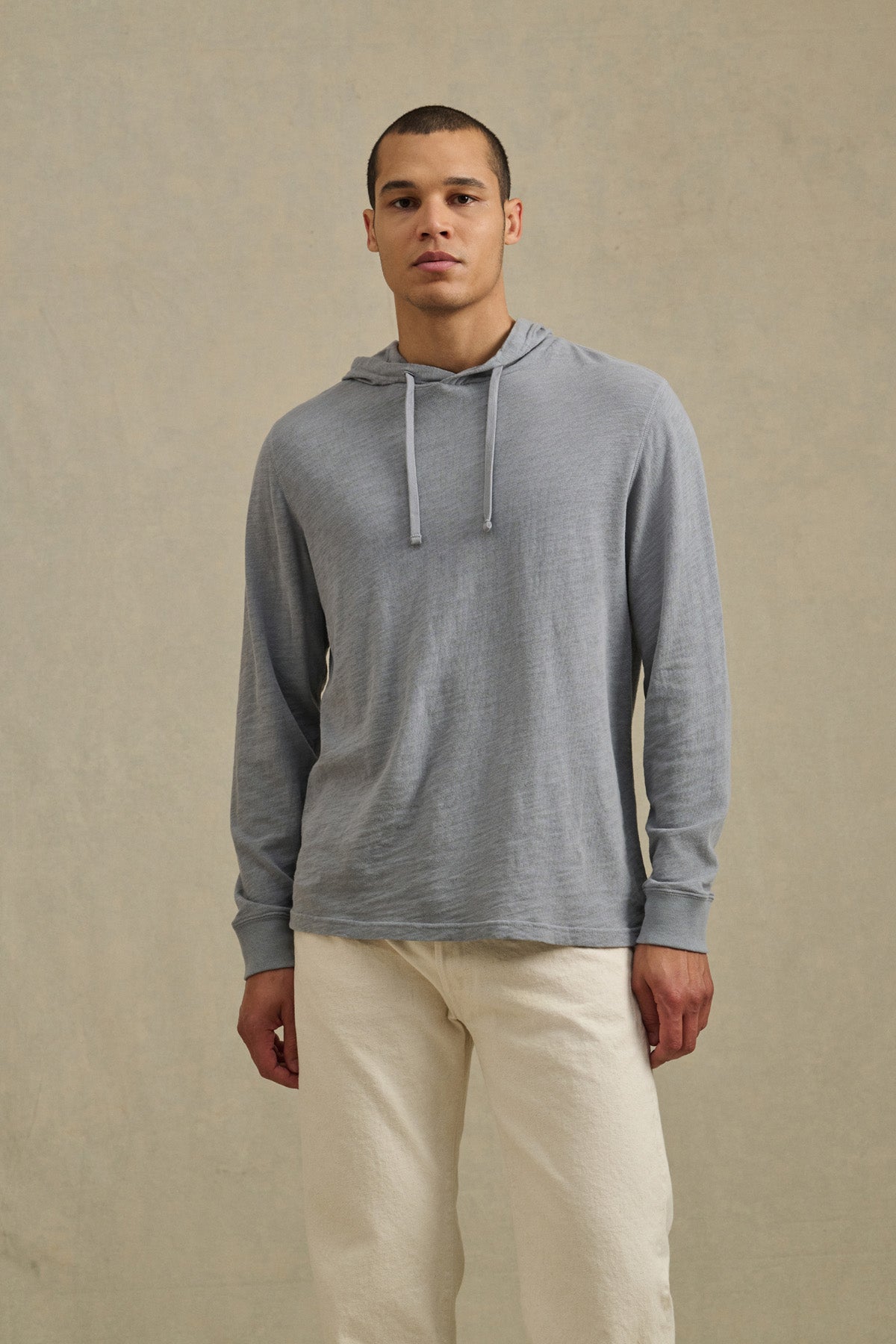 A person wearing the Velvet by Graham & Spencer FRITZ HOODIE in light gray textured cotton with a drawstring hood and off-white pants stands against a plain beige background.-41145282887873