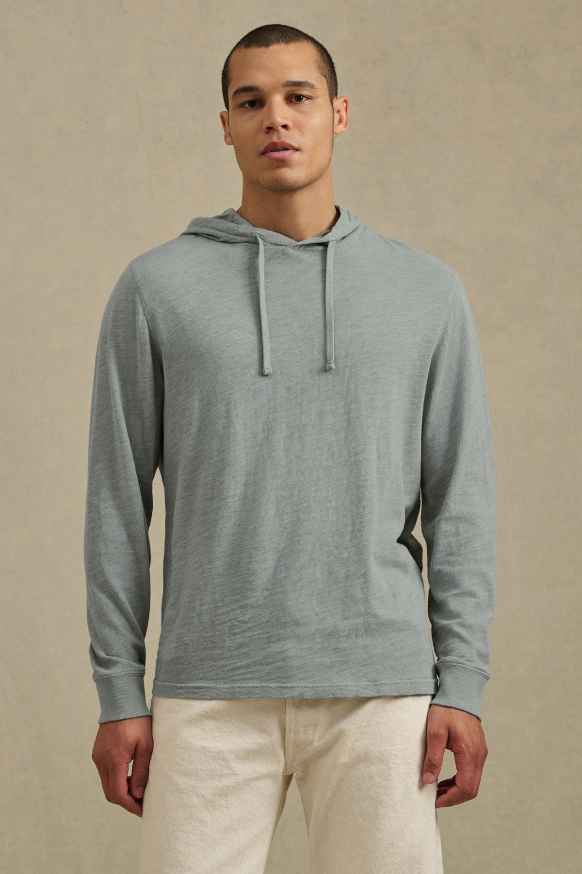 A person stands facing forward against a beige background, wearing the FRITZ HOODIE by Velvet by Graham & Spencer in light gray cotton slub with a drawstring hood, paired with off-white pants for a relaxed, casual look.-41145282855105