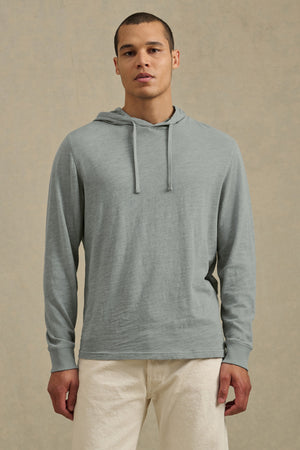 A person stands facing forward against a beige background, wearing the FRITZ HOODIE by Velvet by Graham & Spencer in light gray cotton slub with a drawstring hood, paired with off-white pants for a relaxed, casual look.