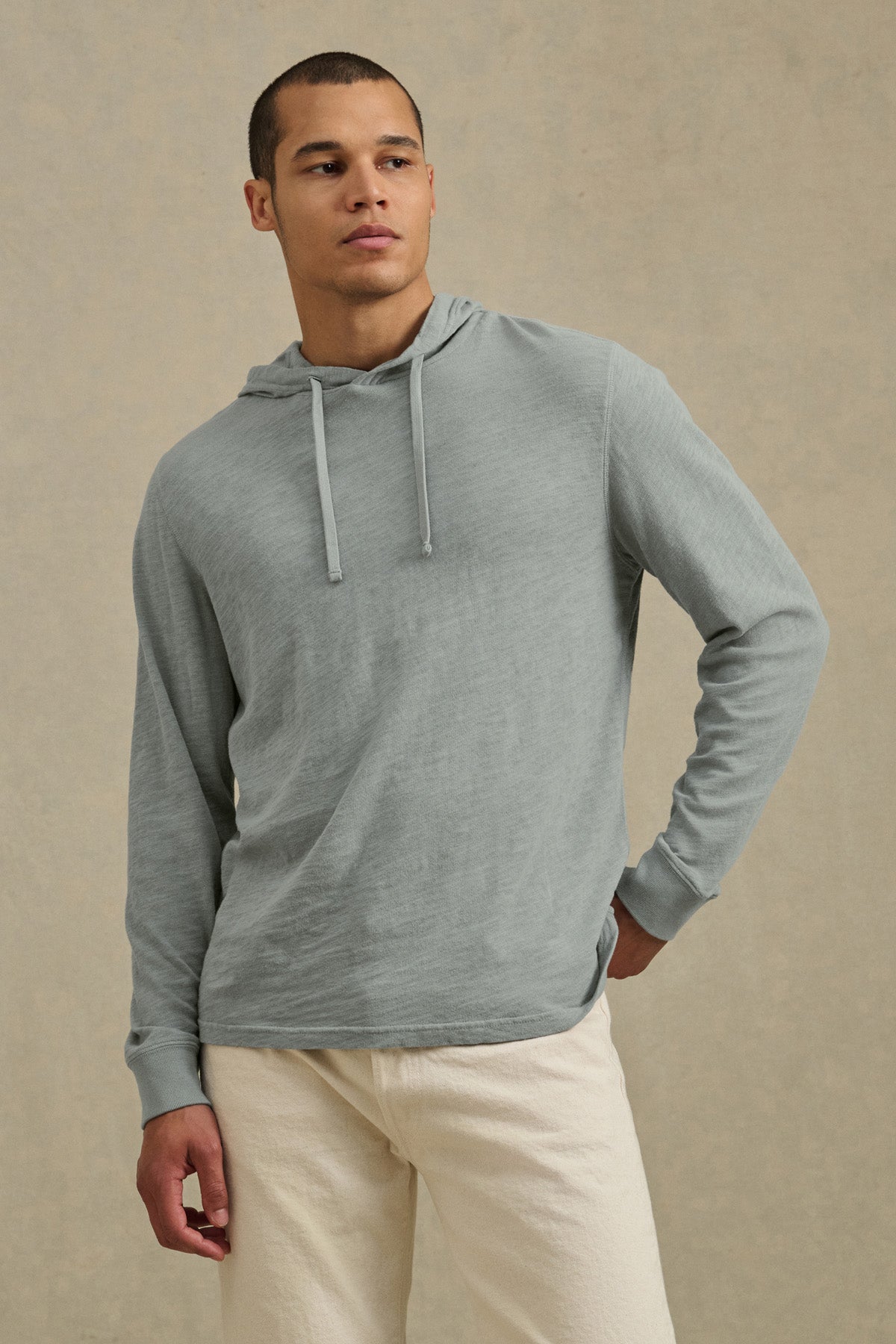 A person wears the FRITZ HOODIE by Velvet by Graham & Spencer in light gray cotton slub with a drawstring hood, paired with off-white pants, standing against a beige background and highlighting relaxed casual style.-41145282822337