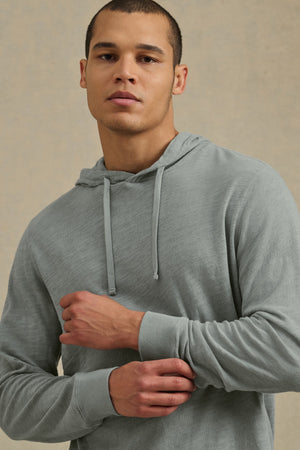 A man wears the FRITZ HOODIE by Velvet by Graham & Spencer—a light gray cotton slub hoodie with a drawstring hood—standing with one arm bent and looking at the camera against a neutral background. Ideal for casual wear.