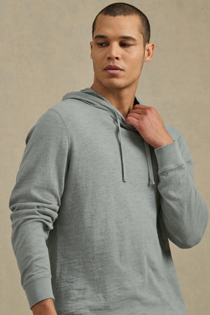 A man wearing the FRITZ HOODIE by Velvet by Graham & Spencer, a light grey cotton slub hoodie with a drawstring hood, looks to his left and holds the collar with his right hand against a plain beige background.