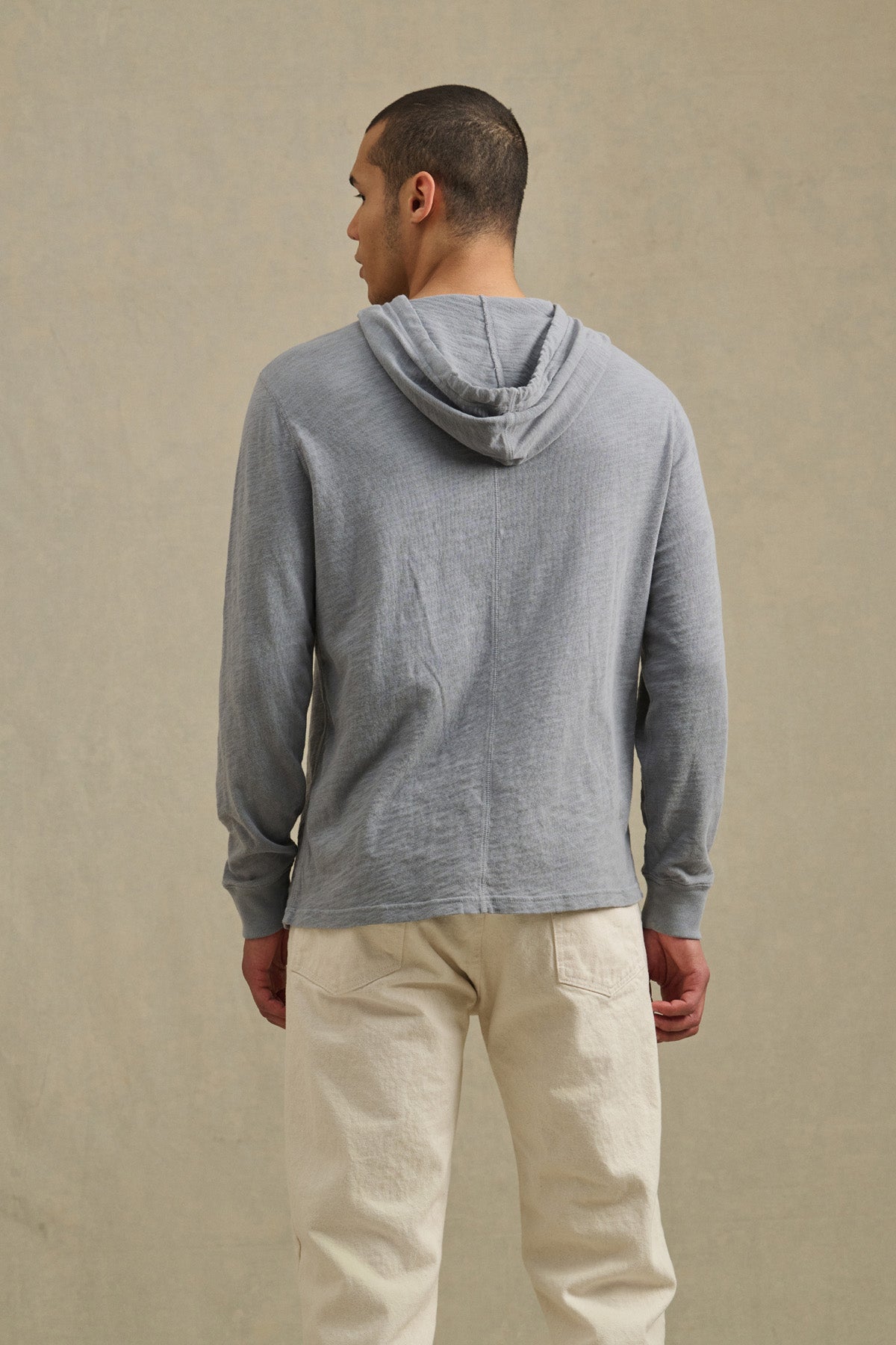 A man faces away, wearing the FRITZ HOODIE by Velvet by Graham & Spencer—a light gray cotton slub hoodie with a drawstring hood—and off-white pants against a neutral background.-41145282724033