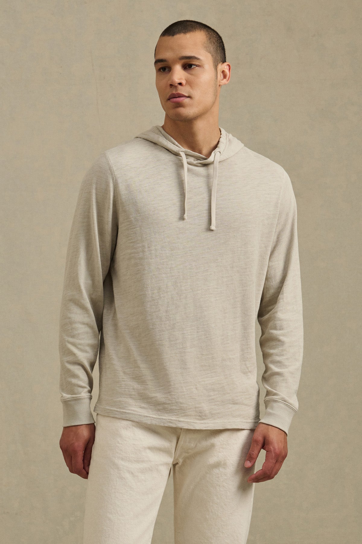 A man stands against a plain background wearing the FRITZ HOODIE by Velvet by Graham & Spencer in light beige cotton slub with matching pants, gazing slightly to the side.