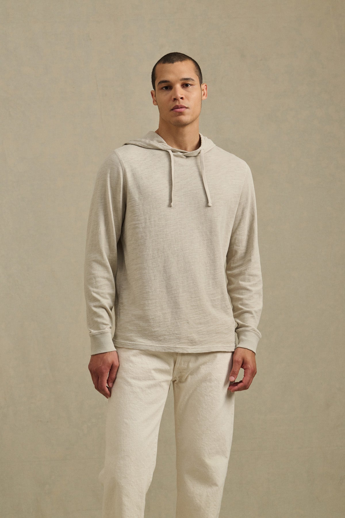 A man stands in front of a neutral background wearing the FRITZ HOODIE by Velvet by Graham & Spencer, a light beige cotton slub hoodie with a drawstring hood, paired with off-white pants.-41104745169089