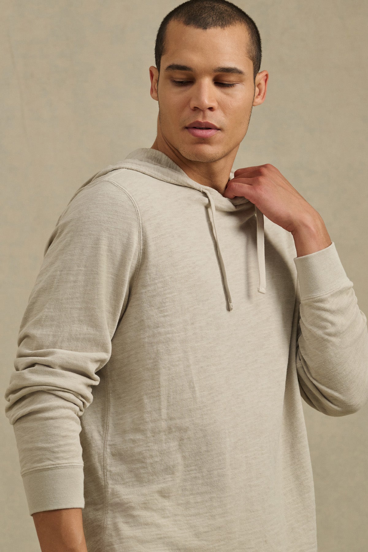 Wearing the FRITZ HOODIE by Velvet by Graham & Spencer—a light beige cotton slub style with ribbed cuffs—a person stands against a plain background, gazing down and to the side while touching the neckline.-41104745136321