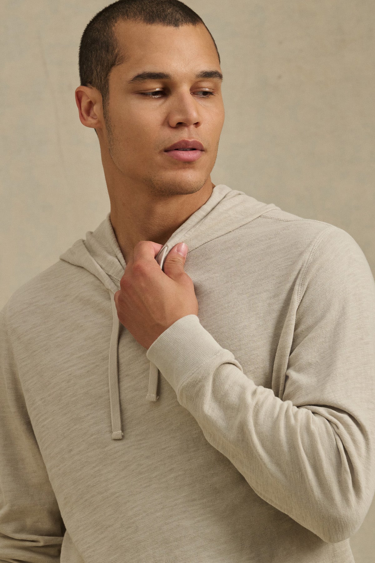 Wearing the FRITZ HOODIE by Velvet by Graham & Spencer in light beige cotton slub, a person looks off to the side while holding the drawstring with one hand.