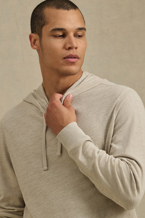 Wearing the FRITZ HOODIE by Velvet by Graham & Spencer in light beige cotton slub, a person looks off to the side while holding the drawstring with one hand.