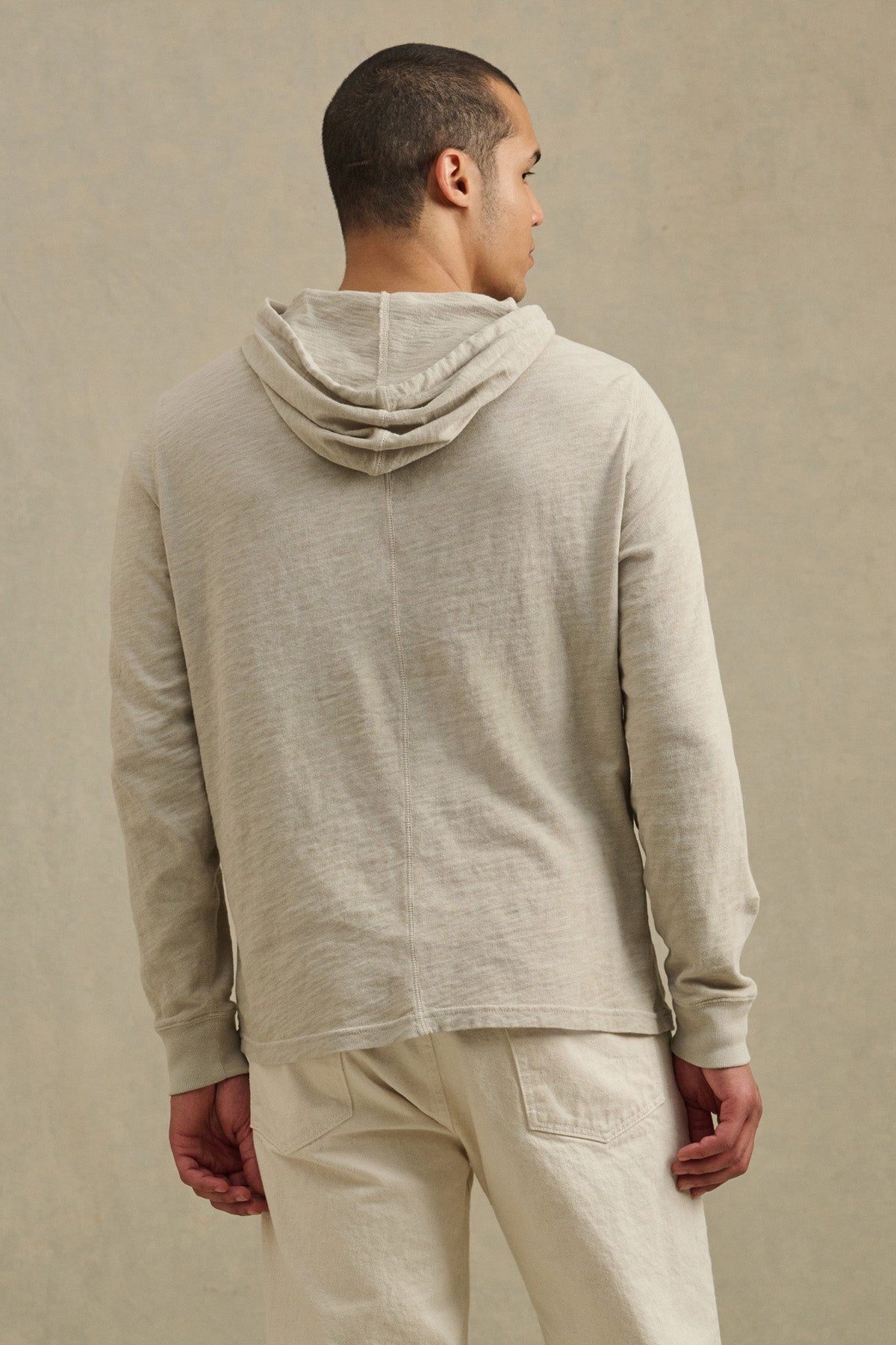 A man faces away from the camera, wearing the FRITZ HOODIE by Velvet by Graham & Spencer in light beige cotton slub, with matching pants, against a plain neutral background.-41104745070785
