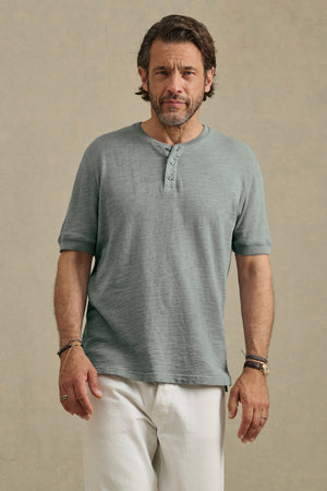 A man with short brown hair and a beard wears the Velvet by Graham & Spencer JENKINS HENLEY in light green and white pants, standing against a neutral background.