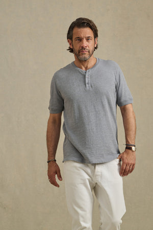 A man with short brown hair and a beard wears the JENKINS HENLEY by Velvet by Graham & Spencer in gray with white pants, standing against a plain beige background.