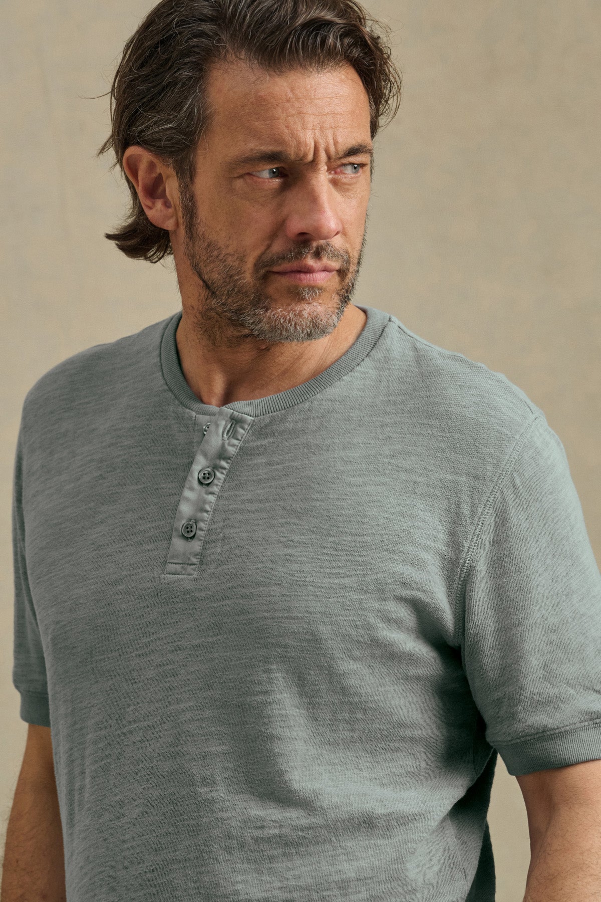 A man with medium-length brown hair and a beard models the JENKINS HENLEY by Velvet by Graham & Spencer, a gray cotton slub top, as he looks to his left against a plain background.-41145292685505