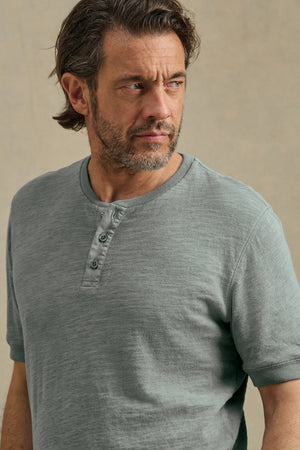 A man with medium-length brown hair and a beard models the JENKINS HENLEY by Velvet by Graham & Spencer, a gray cotton slub top, as he looks to his left against a plain background.