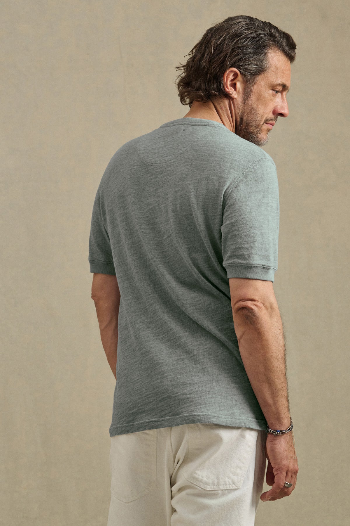 A man with short brown hair wears the JENKINS HENLEY by Velvet by Graham & Spencer in light green and white pants, standing with his back to the camera against a neutral background, embodying easygoing style.-41145292652737