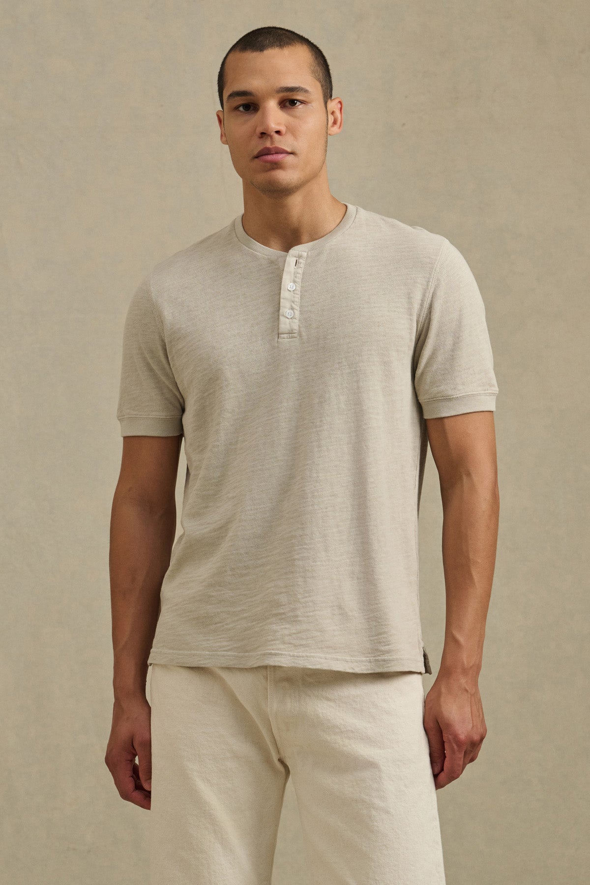 A man faces forward in a Velvet by Graham & Spencer JENKINS HENLEY and cream pants against a plain background, highlighting effortless casual style.