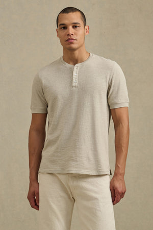 A man faces forward in a Velvet by Graham & Spencer JENKINS HENLEY and cream pants against a plain background, highlighting effortless casual style.
