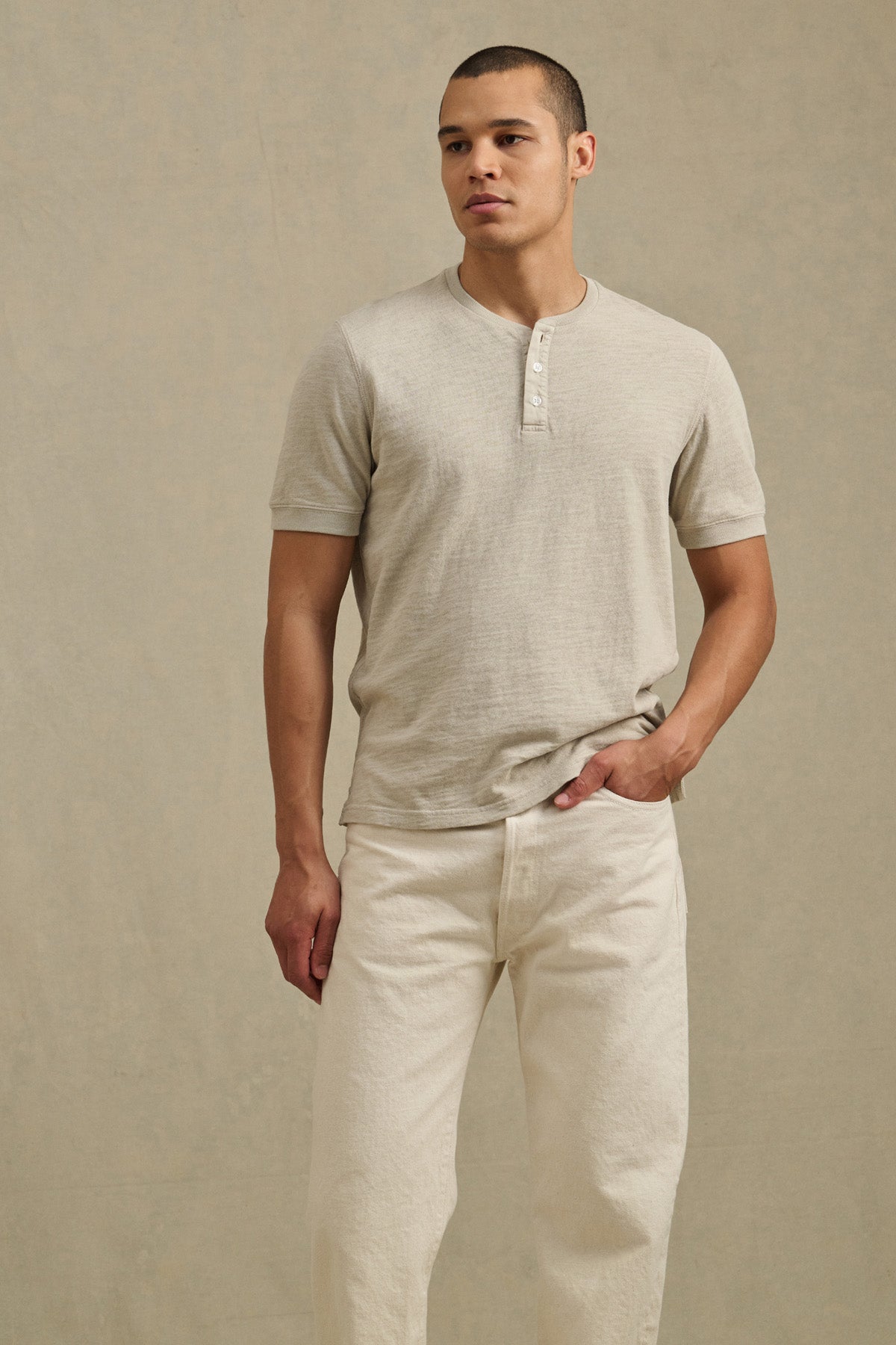 A man models the JENKINS HENLEY by Velvet by Graham & Spencer—a light beige cotton slub short-sleeve shirt—paired with off-white pants, standing against a plain beige background for a laid-back, casual look.-41104761323713