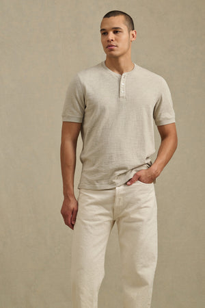 A man models the JENKINS HENLEY by Velvet by Graham & Spencer—a light beige cotton slub short-sleeve shirt—paired with off-white pants, standing against a plain beige background for a laid-back, casual look.