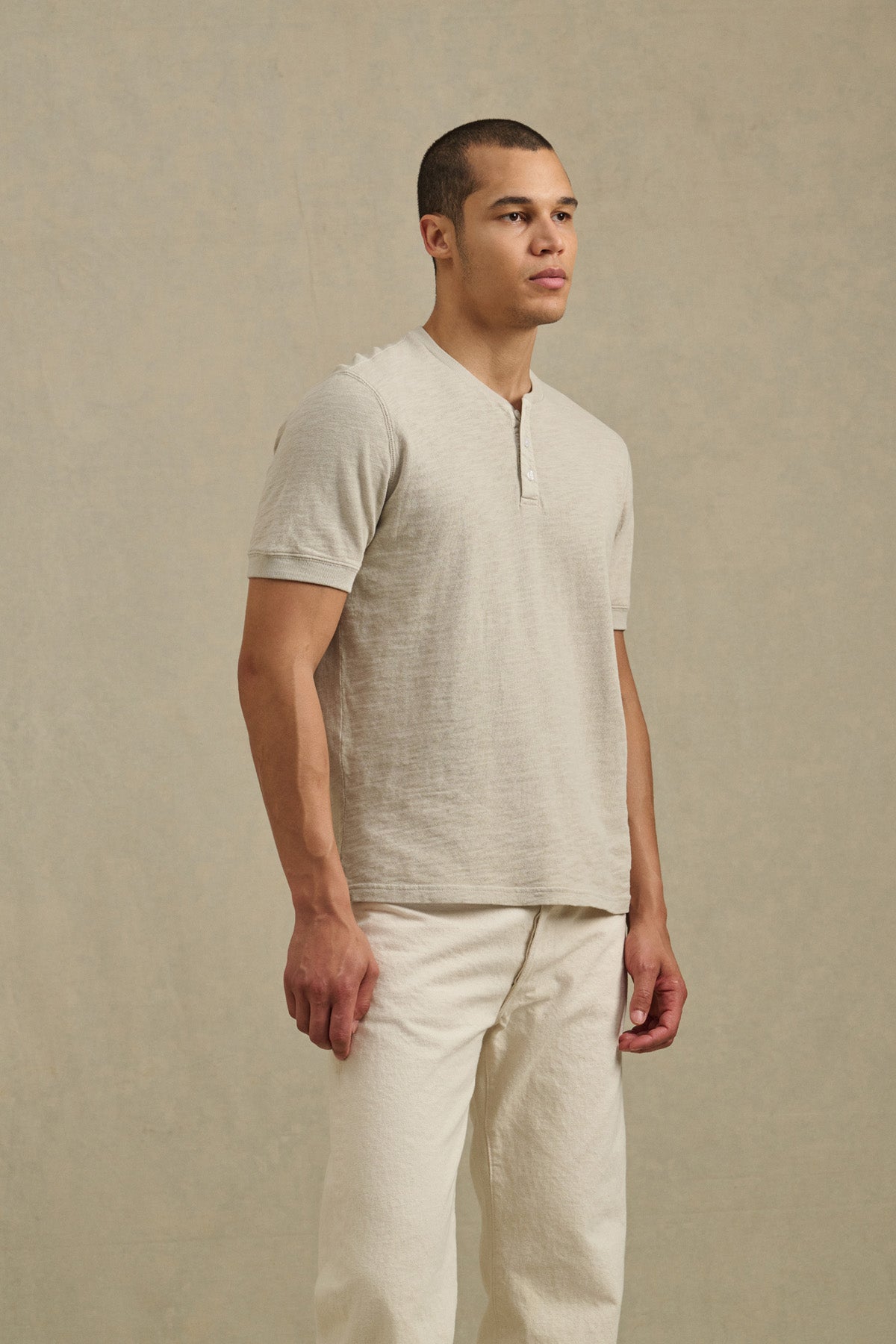 A man stands against a neutral background wearing the Velvet by Graham & Spencer JENKINS HENLEY shirt in light beige cotton slub and off-white pants, offering effortless casual style.-41104761290945