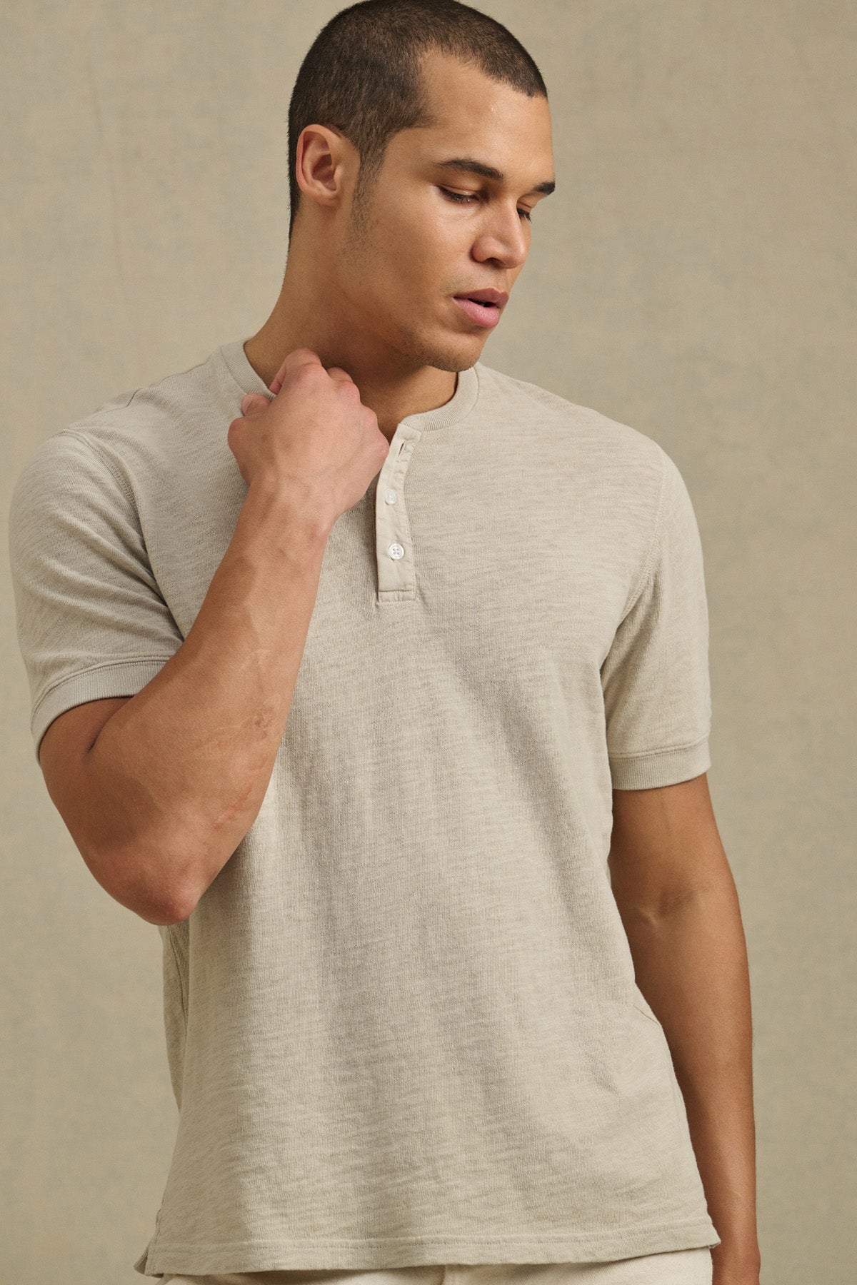 A man wears the JENKINS HENLEY by Velvet by Graham & Spencer, a light beige cotton slub short-sleeve shirt, standing against a plain beige background and looking down with one hand on his neck. Perfect for casual wear.-41104761258177