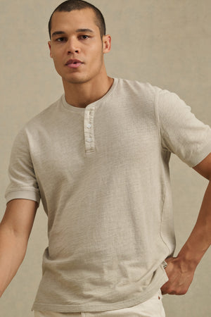 A person models the JENKINS HENLEY by Velvet by Graham & Spencer in light beige cotton, standing against a plain background to showcase effortless casual style.