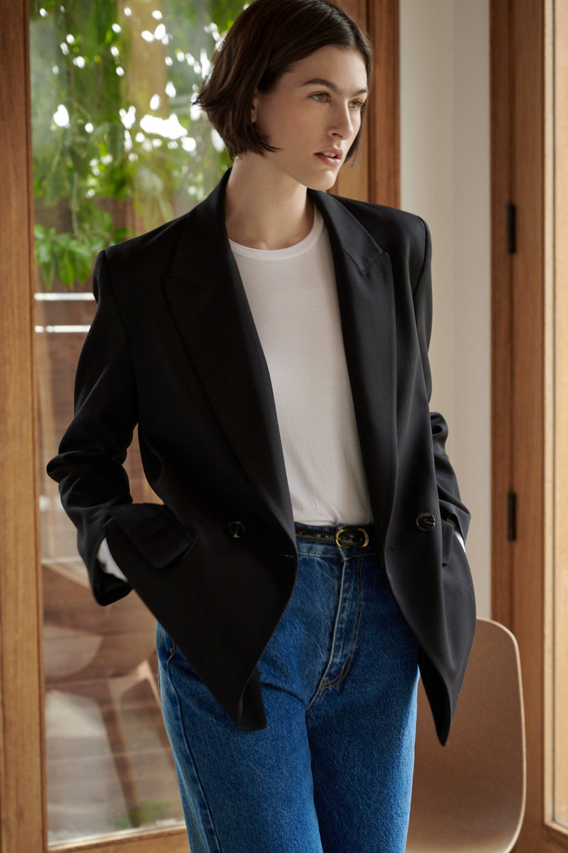 A woman in a Velvet by Jenny Graham FAIRFAX BLAZER and blue jeans standing indoors near a wooden door.