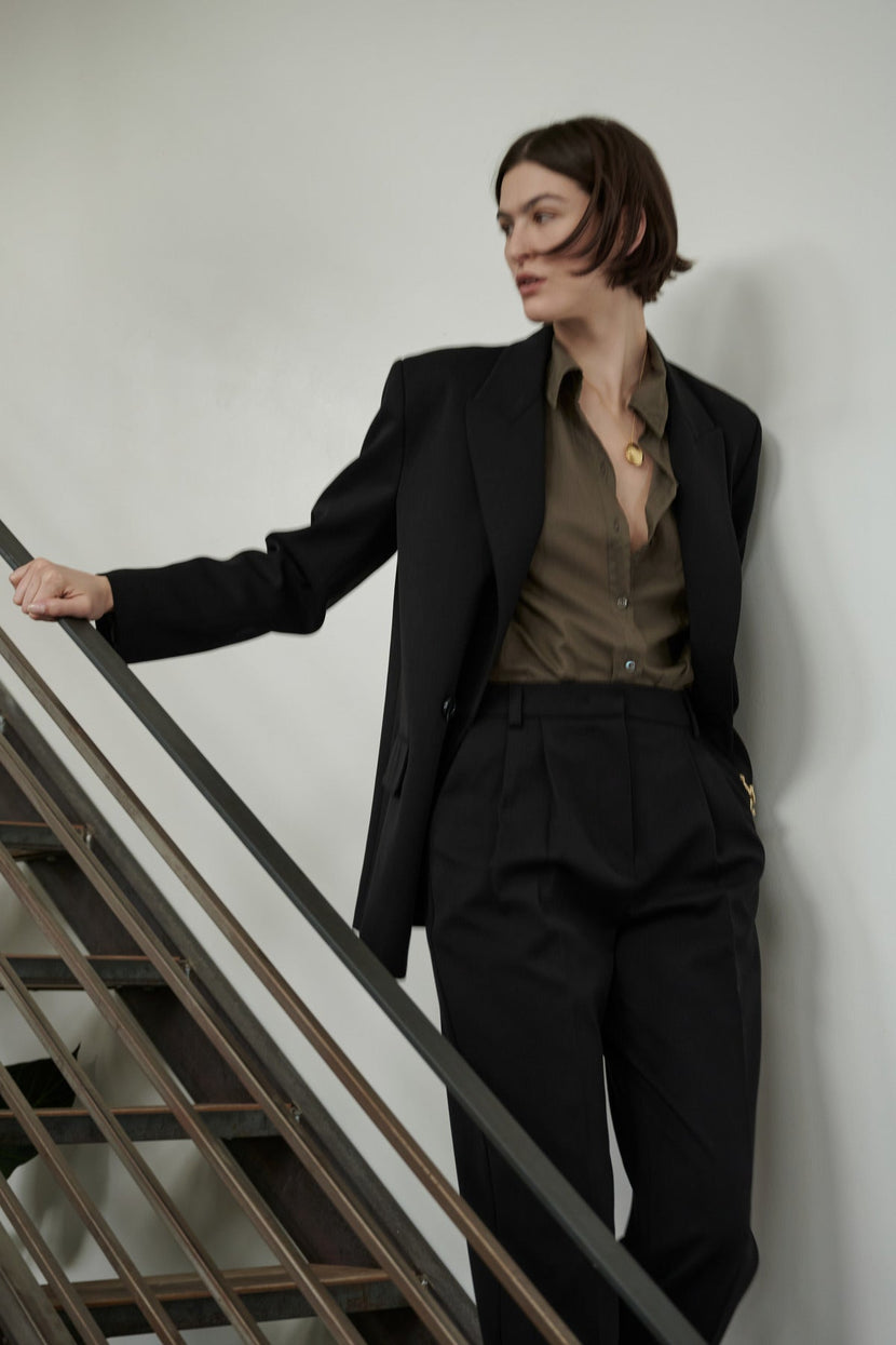 A person in a stylish Velvet by Jenny Graham FAIRFAX BLAZER with structured shoulders and olive shirt posing beside a staircase railing.