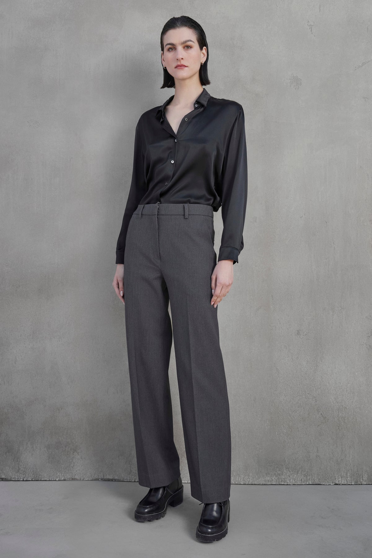   A person stands against a gray textured wall, wearing a black satin long-sleeved shirt, the PRINCE PANT in gray straight-leg suiting fabric by Velvet by Jenny Graham, and black loafers. 