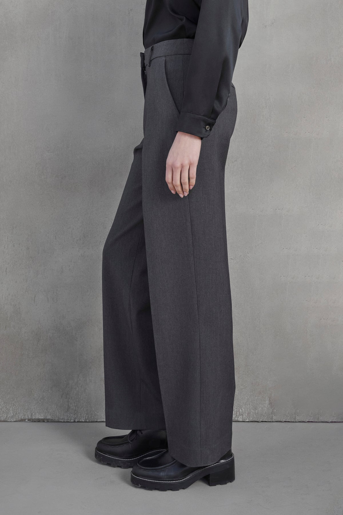 A person standing against a gray background, wearing the high-waisted, wide-leg PRINCE PANT in suiting fabric with pleats from Velvet by Jenny Graham and black shoes.-37659969257665