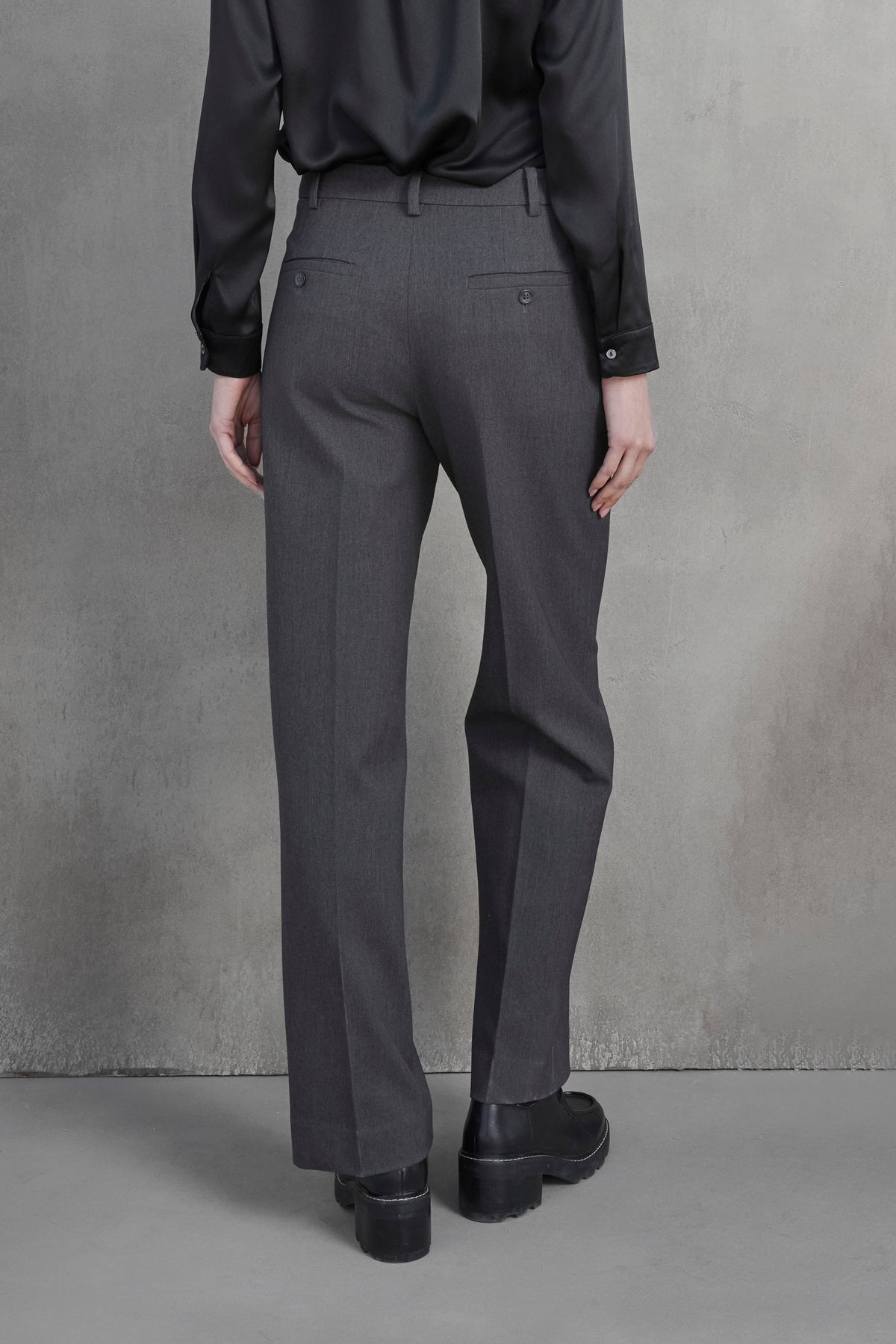   Back view of a person wearing a black long-sleeve shirt, Velvet by Jenny Graham's PRINCE PANT in gray suiting fabric, and black shoes standing against a plain, gray textured wall. 