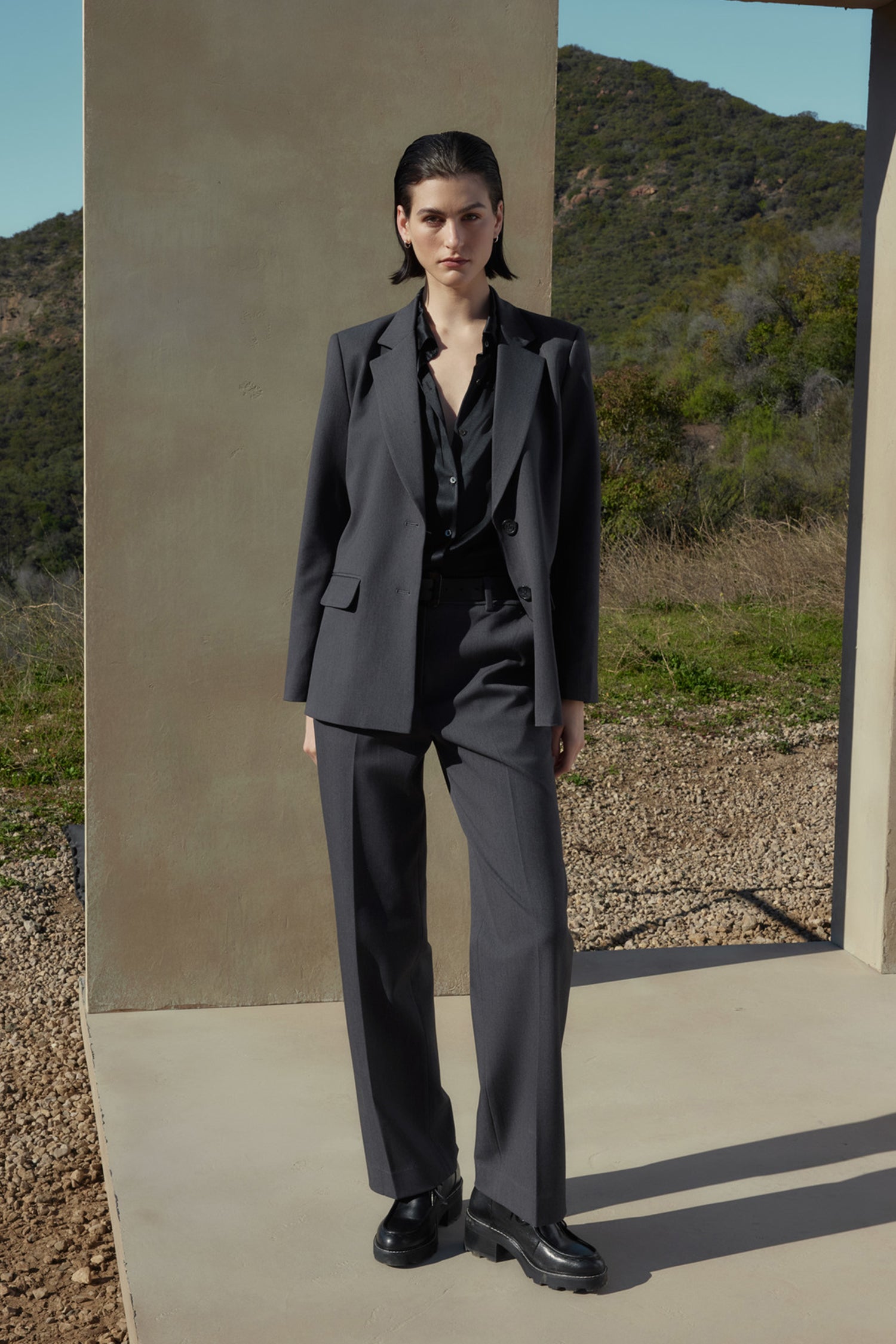   A person with short dark hair wears a grey suit made of suiting fabric and a black shirt, standing on a concrete platform with a mountainous landscape in the background. The PRINCE PANT from Velvet by Jenny Graham completes the polished look. 